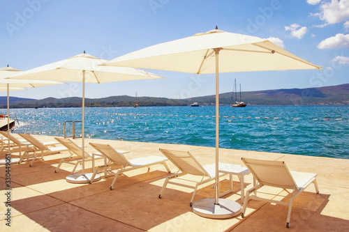 Summer beach vacation. Lounge chairs and white sun umbrellas against blue sky. Montenegro, Adriatic Sea, Bay of Kotor, Tivat city