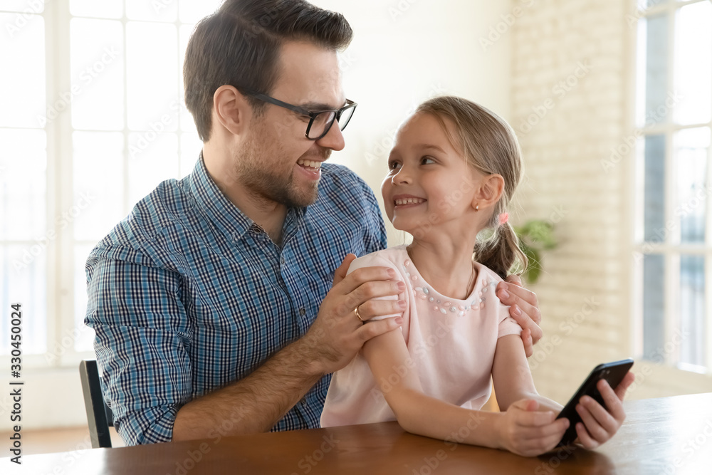 Happy father sitting at table, holding on lap joyful little preschool daughter, using smartphone together. Smiling small child girl playing mobile games online, laughing with dad, relaxing at home. Happy father sitting at table, holding on lap joyful little preschool daughter, using smartphone together. Smiling small child girl playing mobile games online, laughing with dad, relaxing at home.