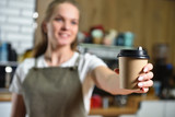 Young girl bartender barista holds out his hand with a paper cup of takeaway coffee. Text space