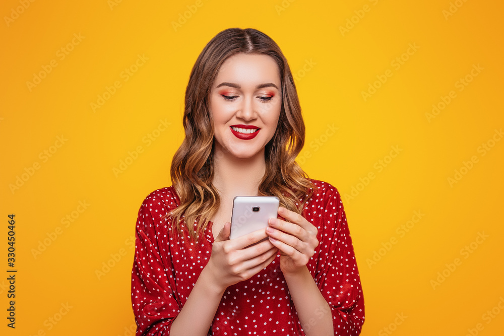 Portrait of a young woman in a red summer dress with red lips chatting by mobile phone isolated over orange wall background. Online orders, purchases, shopping, credit concept Portrait of a young woman in a red summer dress with red lips chatting by mobile phone isolated over orange wall background. Online orders, purchases, shopping, credit concept
