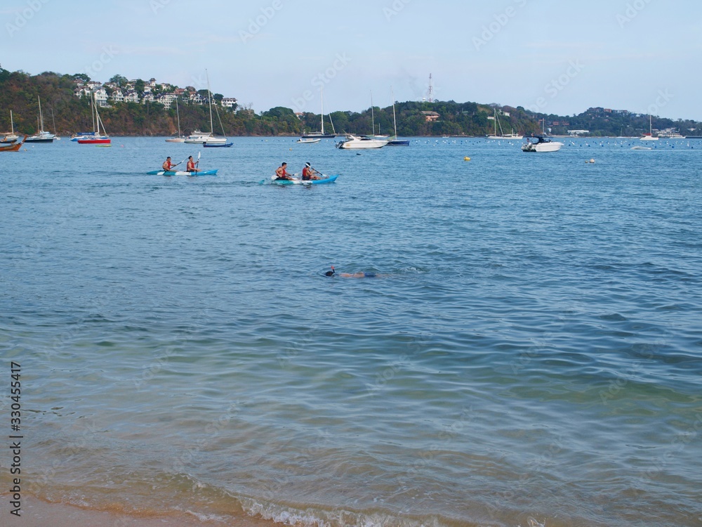 Tropical island, beach, bay with boats and sea water sport activity. Two canoe and paddler moving across water. Snorkeling boy swims under water near kayak.