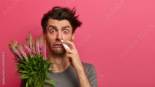 Unhealthy Caucasian man experiences allergy symptoms, caught cold, uses nasal drops, has red nose from sneezing, holds plant allergen, suffers from hay fever, uses effective medicine, stands indoor photo