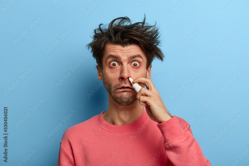 Photo of embarrassed ill man has blocked nose, uses effective medicines, holds bottle of nasal drops for breathing free, wear pink sweater, advertises runny nose remedies. People, cold, treatment Photo of embarrassed ill man has blocked nose, uses effective medicines, holds bottle of nasal drops for breathing free, wear pink sweater, advertises runny nose remedies. People, cold, treatment