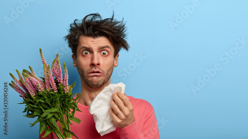 Indoor shot of puzzled man holds handkerhchief, has runny nose and other symptoms of allergy, allergic reaction on plant, has unbelievable gaze at camea, cannot stop sneezing, isolated on blue wall photo
