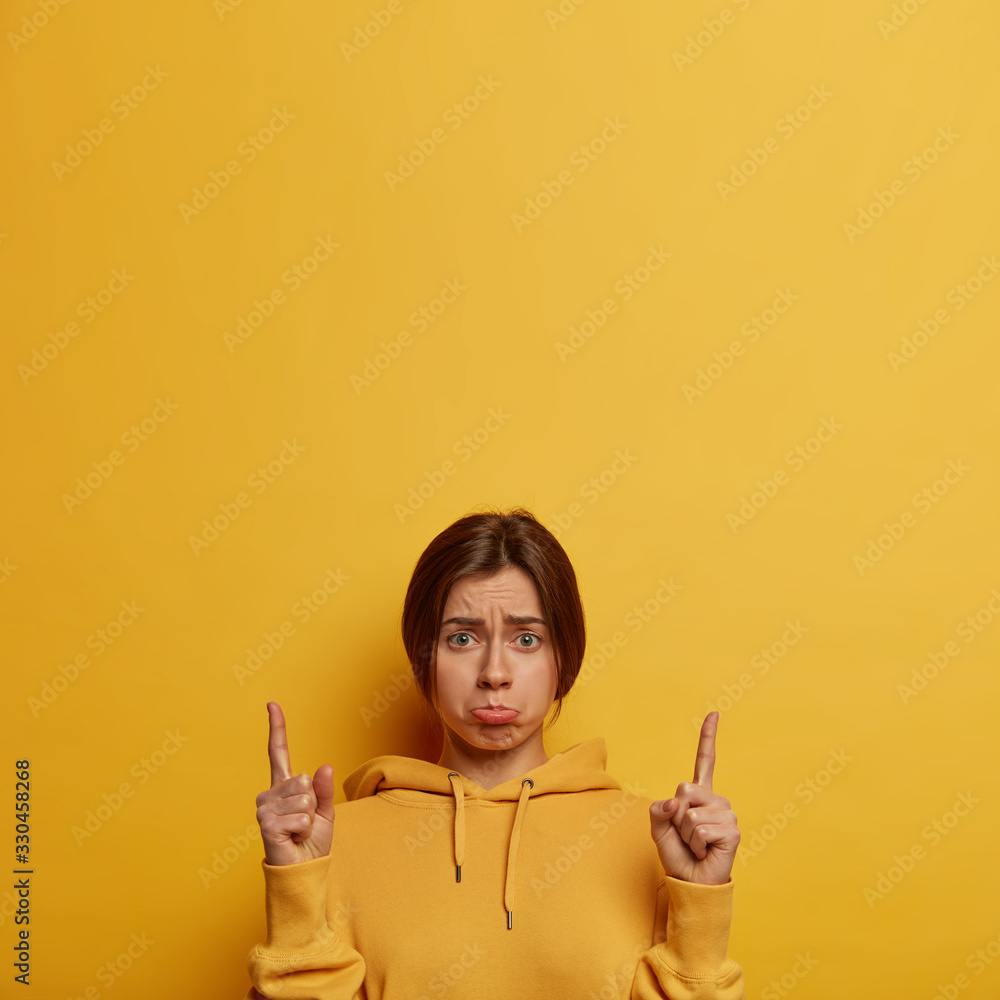 Obraz premium Upset depressed European woman waits for explanation, looks sadly at camera, points fingers up, demonstrates something unpleasant upwards, purses lips, wears hoodie, models over yellow studio wall