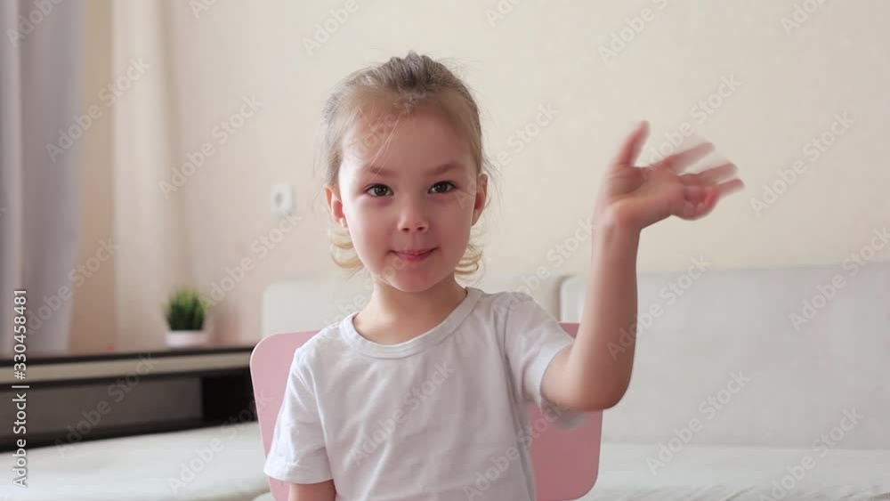 Four years little girl with pretty face smiling looking at camera at home sitting on chair near sofa, talking to webcam making online video call or recording vlog, waving hand Four years little girl with pretty face smiling looking at camera at home sitting on chair near sofa, talking to webcam making online video call or recording vlog, waving hand