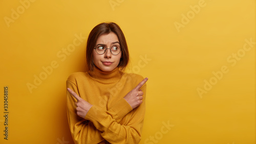 Doubtful Caucasian woman points sideways, tries to choose between two items, asks advice, keeps arms crossed over body, dressed in casual wear, isolated over yellow background, advertises products