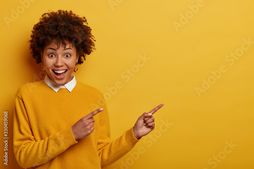 Upbeat positive young woman with Afro hair shows something awesome, attracts your attention to discounts, wears yellow clothes, gestures indoor, involved in advertising campaign, promots item