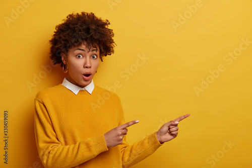 Amazed speechles young female model stares with shocked expression and points aside, tells about awesome discounts, wears casual yellow sweater, poses indoor, witensses something very astounding photo