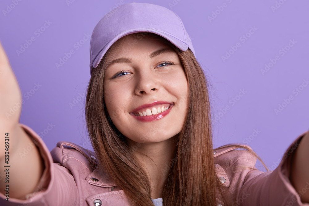 Close up portrait of cheerful tender adorable female holding photocamera, making selfie, looking directly at camera, wearing pink cap, having pleasant facial expression. Youth and tech concept. Close up portrait of cheerful tender adorable female holding photocamera, making selfie, looking directly at camera, wearing pink cap, having pleasant facial expression. Youth and tech concept.