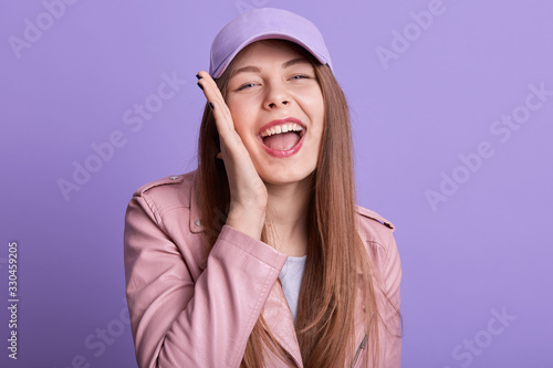 Close up portrait of cheerful sincere magnetic posing isolated over lilac background in studio, opening mouth widely, putting one hand on cheeck, laughing, having fun, feeling good. Emotions concept.