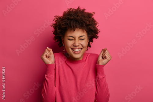 Positive young Afro American woman feels very happy, closes eyes and clenches fists with triumph, feels success to finish project work, wears pink jumper, models indoor gestures actively has good luck