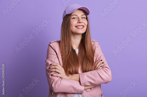 Portrait of positive adorable attractive cute female standing isolated over lilac background in studio, having folded arms, wearing cap and leather jacket, having beautiful smile. Youth concept.