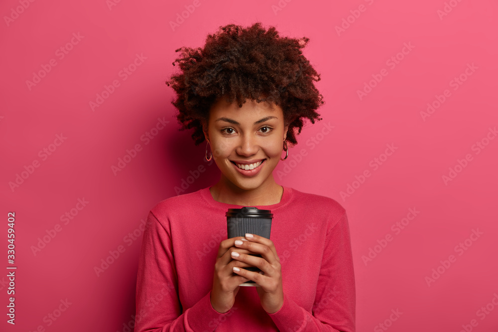 Smiling positive girl holds paper cup of caffeine beverage, looks happily at camera, poses indoor, isolated on pink background, expresses happy feelings. People, drinking, lifestyle concept. Smiling positive girl holds paper cup of caffeine beverage, looks happily at camera, poses indoor, isolated on pink background, expresses happy feelings. People, drinking, lifestyle concept.