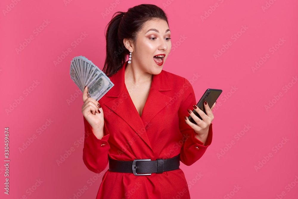 Naklejka premium Indoor image of busy emotional charming young female opening mouth widely, holding fan of money and smartphone in both hands, looking at device screen with surprise. People and money concept.