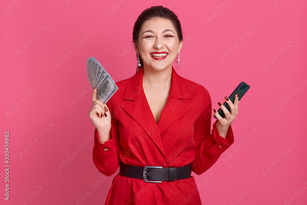 Picture of cheerful happy attractive female smiling sincerely, having delightful facial expression, holding money and smartphone in both hands, wearing red jacket and black belt. Emotions concept. Picture of cheerful happy attractive female smiling sincerely, having delightful facial expression, holding money and smartphone in both hands, wearing red jacket and black belt. Emotions concept.