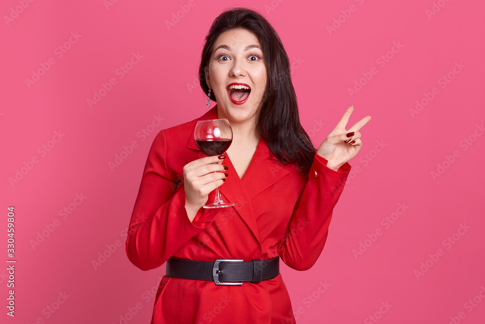 Horizontal shot of young celebrating woman red dress . Beautiful model posing isolated over pink background holding wine glass and showing v sign, screaming something, posing with widely opened mouth. Horizontal shot of young celebrating woman red dress . Beautiful model posing isolated over pink background holding wine glass and showing v sign, screaming something, posing with widely opened mouth.