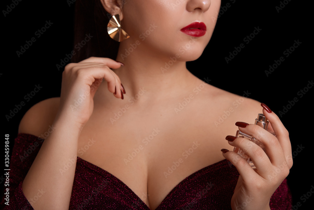 Perfume bottles in hands of beautiful girl oi studio isolated over black background, part of face of young woman with aroma, concept of care and cosmetics, female wearing elegant dress and earrings.