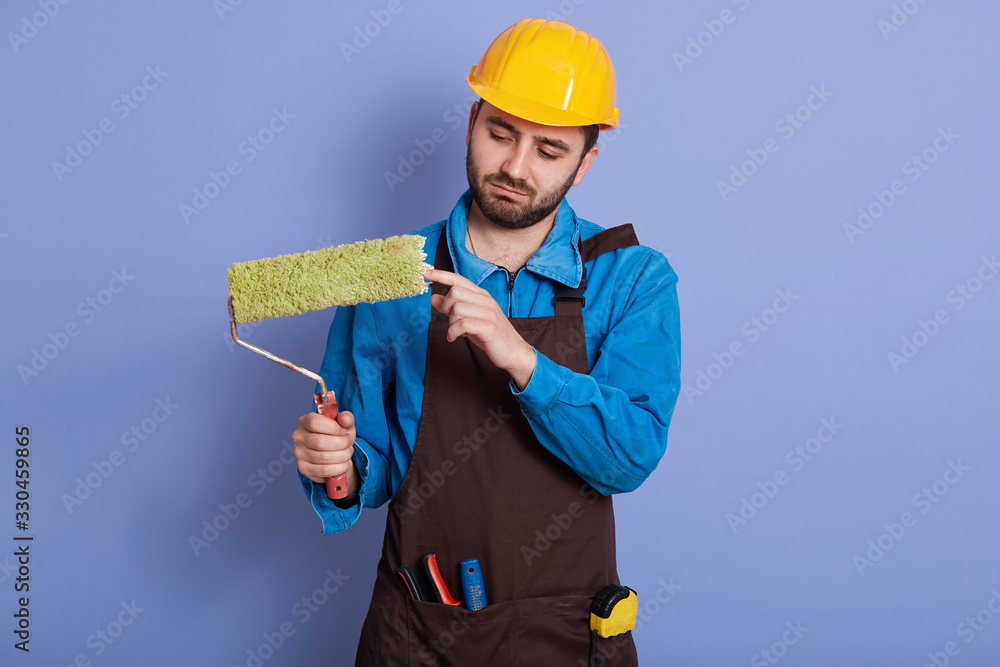 Horizontal picture of attentive hard working young repairman holding roller in one hand, wearing blue overall, brown apron and yellow helmet, having equipment. People and working process concept. Horizontal picture of attentive hard working young repairman holding roller in one hand, wearing blue overall, brown apron and yellow helmet, having equipment. People and working process concept.