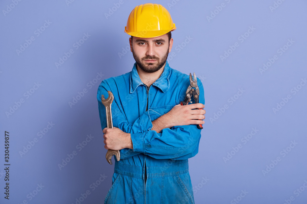 Indoor portrait of confident handsome bearded worker standing isolated over blue background in studio, holding pliers and wrench in both hands, being ready to work. People and engineering concept.