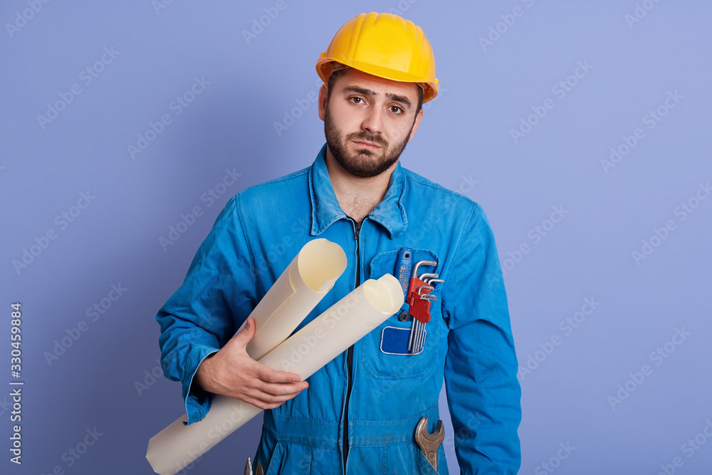 Image of tired confused upset worker having working day, holding plans of projects, having equipment, posing isolated over blue background in studio, wearing uniform. People and work concept.