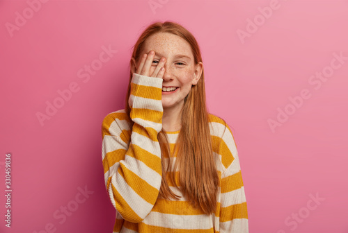 Fotografi Carefree optimistic ginger girl keeps palm on face, smiles toothily, giggles as hears jokes, has long foxy hair, freckled face, wears loose jumper, isolated on pink wall