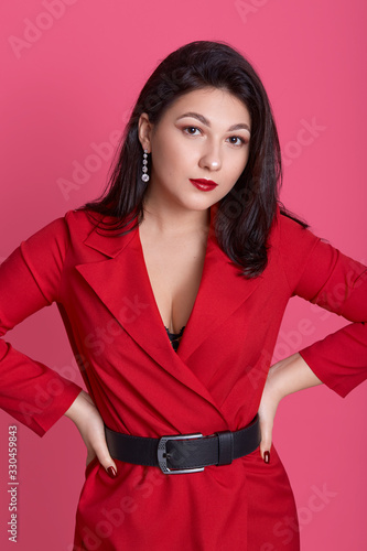 Close up portrsit of beautiful young woman wearing elegant redd dress with black belt posing with hands on hips and looking at camera isolated over pink studio background, female looks confident. photo