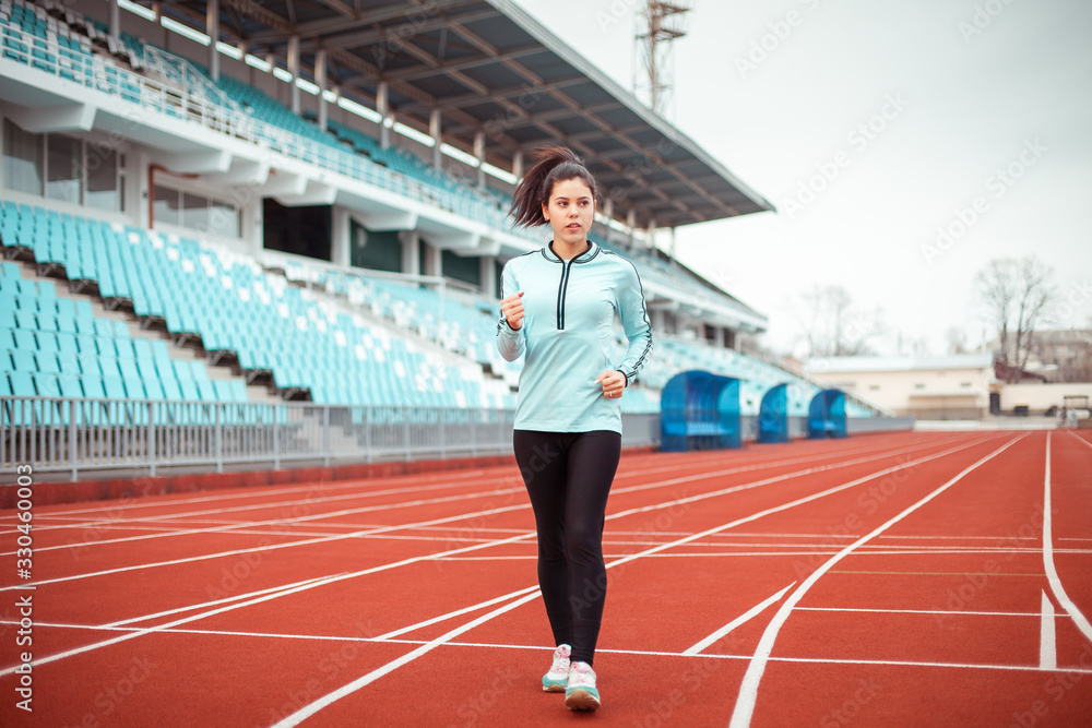Beautiful girl goes in for sports in the stadium. The girl trains and does exercises in a blue sports uniform on a brown track.