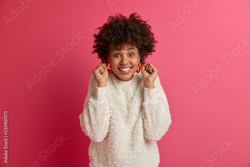 Foto Overjoyed curly woman smiles broadly, makes fist bump, anticipates great news, expresses positive emotions, dressed in casual wear, cheers good news, isolated over pink background