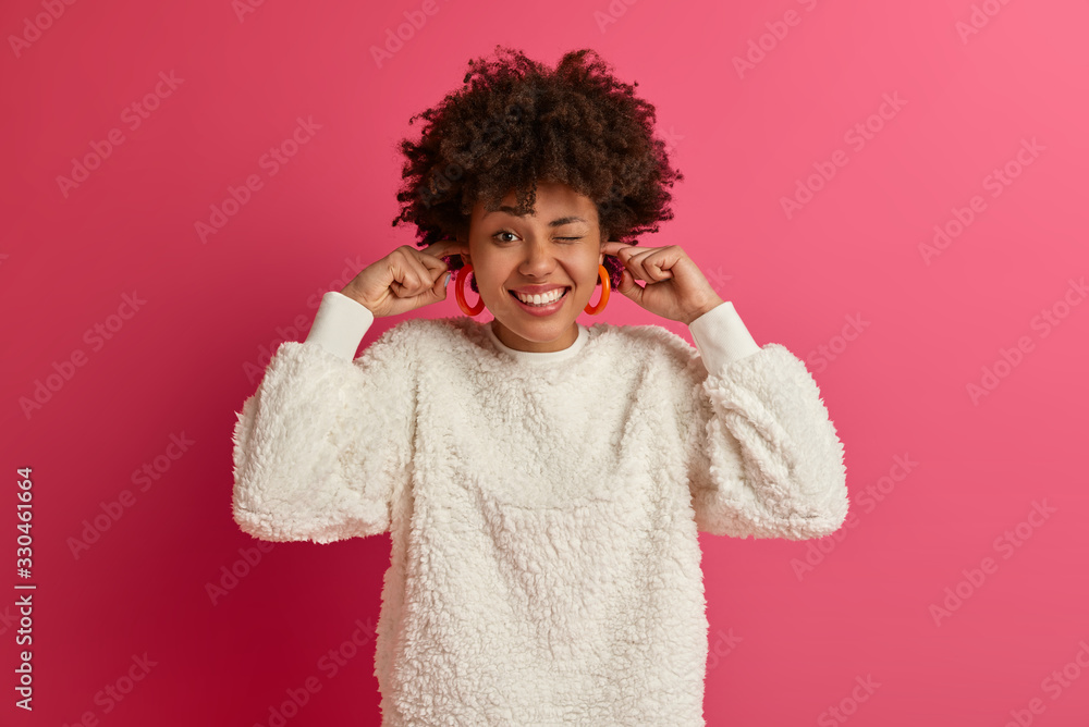 Joyful ethnic woman winks eye, smiles broadly, plugs fingers into ear