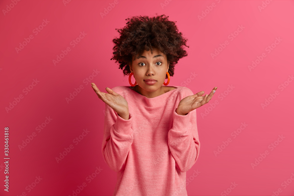 Photo of doubtful Afro American lady shrugs shoulders, makes decision, faces dilemma, feels apathy, hesitates as makes choice, looks questioned, isolated on pink background. People, attitude