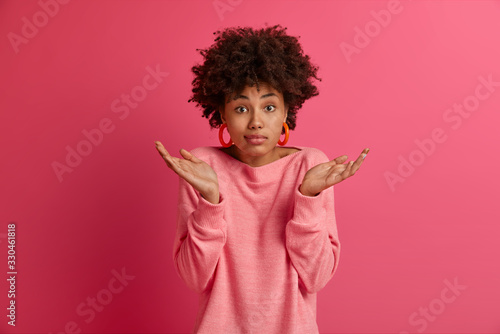 Tableau sur toile Photo of doubtful Afro American lady shrugs shoulders, makes decision, faces dilemma, feels apathy, hesitates as makes choice, looks questioned, isolated on pink background