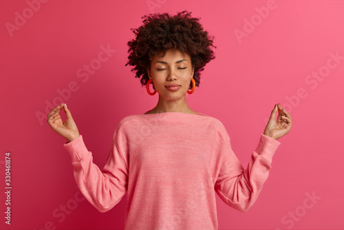 Relaxed curly haired woman meditates and makes yoga exercises, raises hands sideways in lotuse pose, closes eyes and reaches nirvana, wears casual jumper, isolated over bright pink background photo
