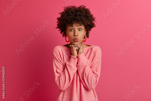 Indoor shot of pretty Afro American woman keeps lips folded  ready to receive kiss  keeps both hands under chin  has surprised expression  isolated on pink background. Human face expressions concept