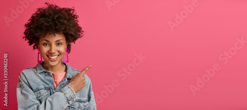 Horizontal shot of pleasant looking smiling female model indicates at copy space, advertises something nice and appealing, gives advice, wears fashionable denim jacket, isolated on pink background