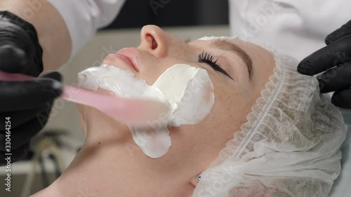 In beauty salon. Cosmetologist putting mask on female faceskin in spa salon. Beautician applying lifting cream using stick, Pretty young woman getting beauty procedure with clay applied with spatula photo