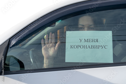 A woman with long black hair is sitting in a car and holding a sign with the inscription in Russian. I have a coronavirus