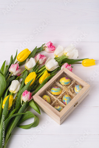 Fototapeta Naklejka Na Ścianę i Meble -  Top view of colorful easter eggs on a bed of straw in a long wooden box on a white wooden table and tulips, place for text