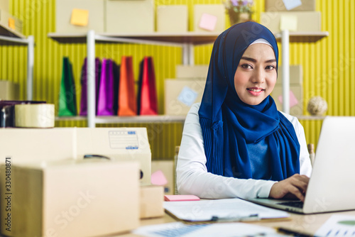 Portrait of smiling beautiful muslim owner asian woman freelancer sme business online shopping working on laptop computer with parcel box on table at home - Business online shipping and delivery