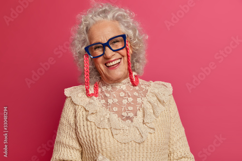 Pleased curly haired mature lady wears transparent glasses and knitted sweater, happy children come to visit her, being in good mood, enjoys life, poses over pink background. People and age concept