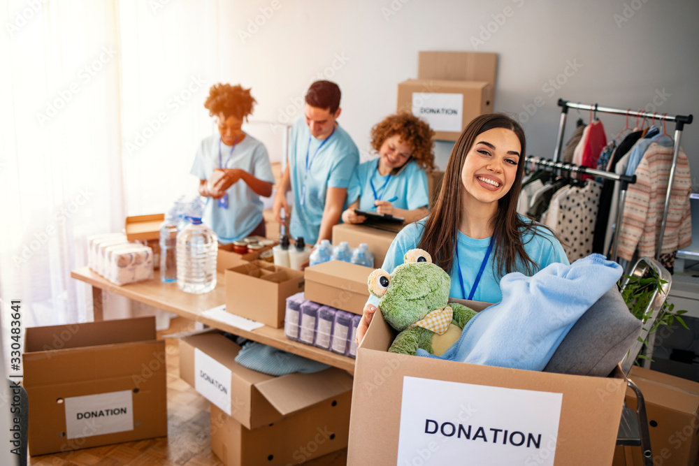 We are successful team of social workers. Woman volunteer holding a donation box and is smiling at the camera. Volunteers are helping people in the background. We are successful team of social workers. Woman volunteer holding a donation box and is smiling at the camera. Volunteers are helping people in the background.