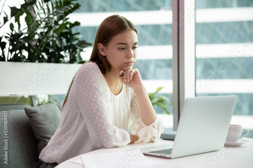 Pensive millennial Caucasian female employee sit at desk working on laptop thinking planning, thoughtful young woman work or study on computer, make notes, take online course or training