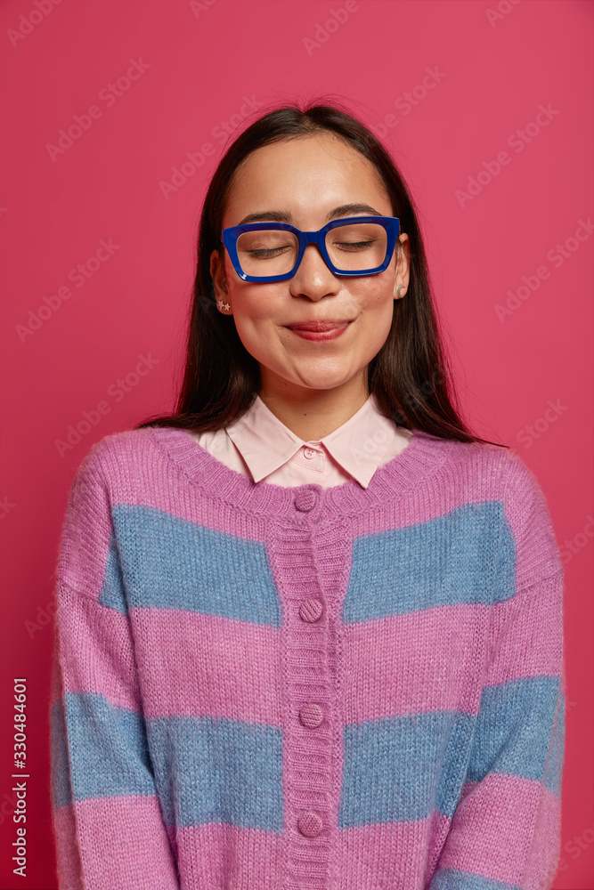 Photo of charming clever female nerd with Asian appearance wears big square glasses striped jumper, keeps eyes shut, feels shy, poses against pink backgroundglad to receive praise for well done work Photo of charming clever female nerd with Asian appearance wears big square glasses striped jumper, keeps eyes shut, feels shy, poses against pink backgroundglad to receive praise for well done work