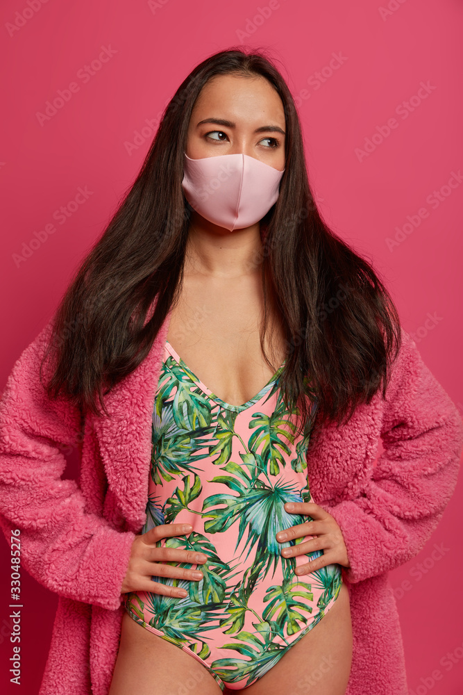 Vertical shot of thoughtful woman wears swimsuit and domestic robe, has spoiled travel vacation because of coronavirus outbreak, protects face with mask, thinks abot risk of epidemic disease Vertical shot of thoughtful woman wears swimsuit and domestic robe, has spoiled travel vacation because of coronavirus outbreak, protects face with mask, thinks abot risk of epidemic disease