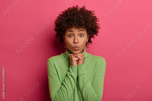 Good looking young ethnic woman keeps lips rounded  keeps hands under chin  looks directly at camera  has widely opened eyes  dressed in casual wear  poses against pink background  waits for kiss