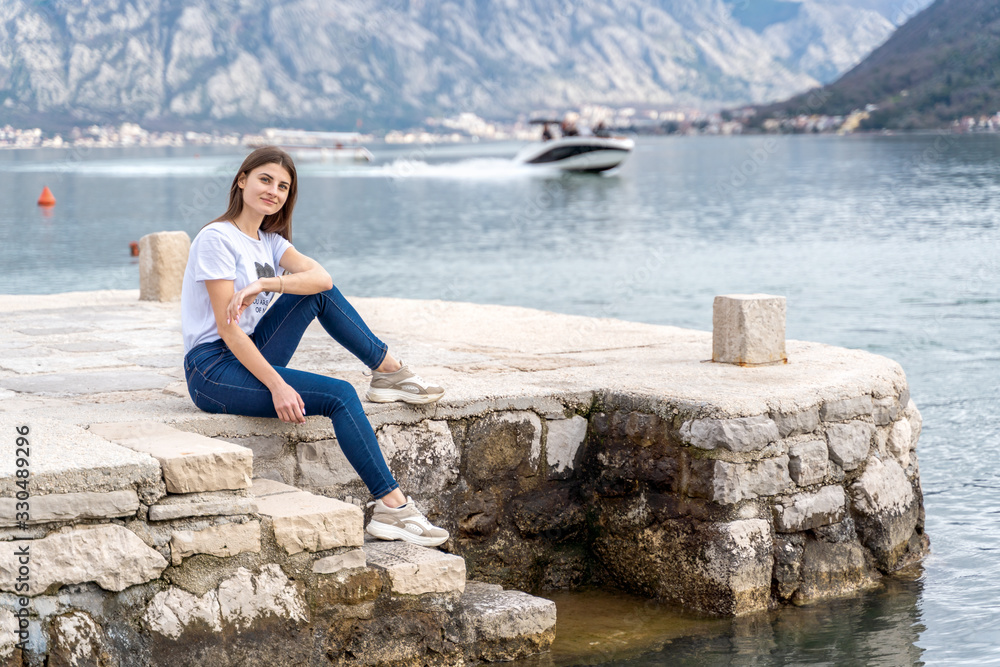 Woman in orange hoodie stands by the sea in city Perast on the background in Montenegro