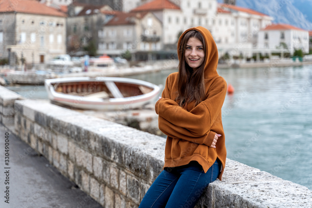 Woman in orange hoodie stands by the sea in city Perast on the background in Montenegro Woman in orange hoodie stands by the sea in city Perast on the background in Montenegro