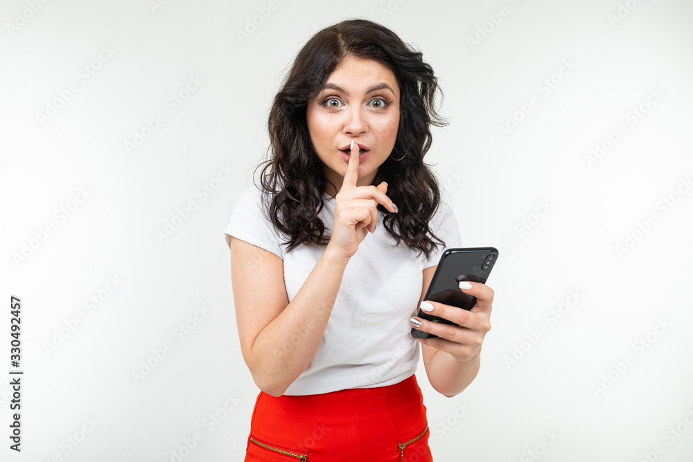 girl keeps a secret while holding a finger at her mouth and a smartphone in her hand on a white background girl keeps a secret while holding a finger at her mouth and a smartphone in her hand on a white background
