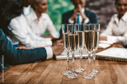 Five Glasses With Alkohol Are Lokated On Wooden Table In Office. Selective Focus On Wineglasses With Champagne, Meeting Of Business People on Blured Background, Toned Image photo