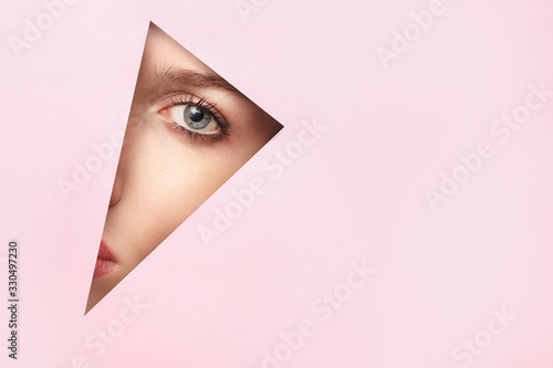 Face of a young beautiful woman with make-up peers into a hole in pink paper.